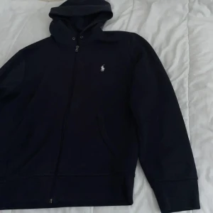 Svart hoodie från Ralph Lauren - Svart hoodie från Ralph Lauren med dragkedja framtill och klassisk broderad logga på bröstet. Tröjan har huva, långa ärmar och två fickor framtill. Perfekt för en chill och stilren look.