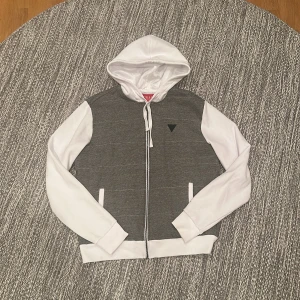Guess Hoodie - Snygg hoodie från Guess med vit huva och ärmar samt gråsvart randig kropp. Dragkedja framtill, två fickor och klassisk Guess-logga på bröstet. Perfekt för en chill och stilren look.