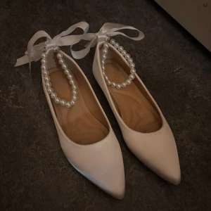 Vita ballerinaskor med pärlband och rosett - Snygga vita ballerinaskor med spetsig tå, dekorerade med pärlband runt vristen och breda sidenband att knyta. Skorna har platt sula och är tillverkade i ett glansigt material som ger en elegant look. Perfekta för dig som vill sticka ut med detaljer.