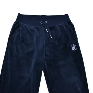 Juicy Couture mörkblå velourbyxor - Mörkblå mjukisbyxor från Juicy Couture i mjuk velour med raka ben, snörning i midjan och fickor. Byxorna har glittriga strasstenar med Juicy-logga på ena benet och juicy loggan på rumpan. Perfekta för en chill och trendig look.