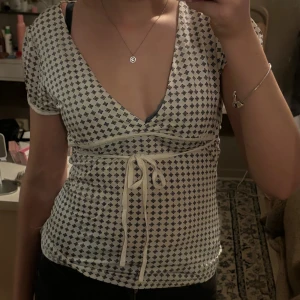 Vit omlottopp med knyt - Supersöt vit omlottopp från Vero Moda med små svartmönstrade detaljer. Toppen har v-ringning, korta ärmar och knytband i midjan som ger en snygg passform. Perfekt till jeans eller kjol för en fräsch look.