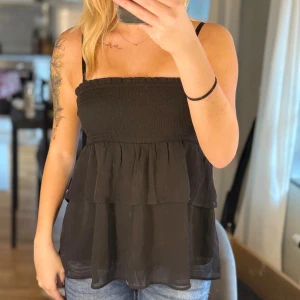 Svart volangtop från Gina Tricot - Säljer en svart ärmlös topp från Gina Tricot med smockad överdel och dubbla volanger nedtill. Toppen har tunna axelband och är tillverkad i ett lätt, luftigt material som ger en flowy känsla. Perfekt att styla med jeans eller kjol.