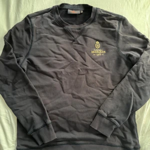 Morris tröja - Snygg mörkgrå sweatshirt från Morris med broderad gul logga och emblem på bröstet. Tröjan har rund hals, ribbade muddar och är tillverkad i mjuk bomull. Perfekt för en chill och stilren look.