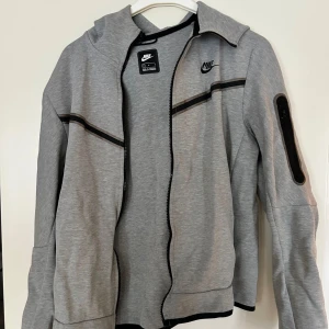 Nike Tech storlek m - Snygg grå vindjacka från Nike med svarta detaljer och dragkedja framtill. Jackan har huva, lång ärm med ficka på ena ärmen och sportig look. Den är lite riven på armbågen, alltså den har tvättats rätt många gånger. Perfekt för dig som gillar streetstyle och vill ha något bekvämt och stilrent. Köptes för 1200kr