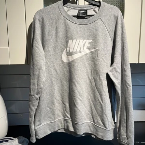 Grå sweatshirt från Nike med logga - Snygg grå sweatshirt från Nike i storlek L med klassisk vit Nike-logga på bröstet. Tröjan har rund hals, långa ärmar och är gjord i mjuk bomullsmix som är perfekt för chill eller träning. Enkel och stilren design som funkar till jeans eller joggers.