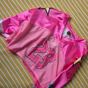 Rosa topp  - Cool rosa topp  med glänsande tyg, mesh framtill och stort nummer 23. Tröjan har lång ärm, V-ringning och dekorativa stjärnor på axlarna. Perfekt för dig som vill sticka ut på match eller i vardagen.