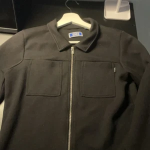 Svart overshirt från Jack & Jones - Svart overshirt från Jack & Jones med två stora bröstfickor och silverfärgad dragkedja framtill. Klassisk krage och stilren design som funkar till många olika outfits. Materialet känns mjukt och har en snygg, enkel look.