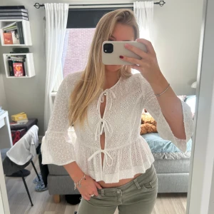 Vit blus med knyt och volang - Superfin vit blus med broderade hålmönster och tre knyt framtill. Blusen har volang nedtill och vida trekvartsärmar. 