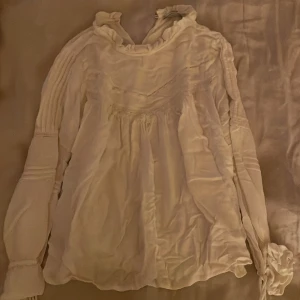 Vit blus med volang från Mango - Supersöt vit blus från Mango med volangdetaljer vid halsen och ärmsluten. Blusen har långa ärmar med snygga rynkningar och en luftig, romantisk passform. Perfekt för dig som gillar en feminin och trendig stil.