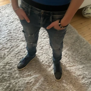 Blå slitna jeans med skinny fit - Säljer ett par blå jeans med skinny passform och slitna detaljer på benen. Jeansen har klassisk femficksdesign, dragkedja och knapp i midjan. Materialet är jeans och färgen är en tvättad blå nyans med ljusare partier. Perfekta för en avslappnad och trendig look.