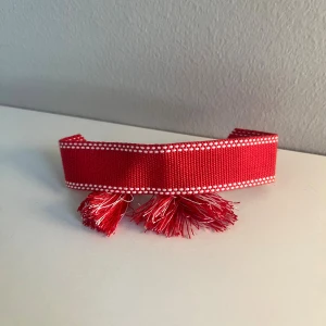 Rött armband nytt - Säljer ett unikt rött armband med vita prickade kanter och två dekorativa tofsar framtill. Armband är brett och har en cool textil känsla, perfekt för att ge outfiten en färgklick och personlig touch.