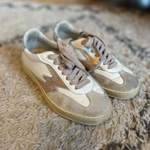 Moaconcept skor!! - Snygga sneakers från Moaconcept i vitt och beige med metallic guld-blixt på sidan. Skorna har rund tå, platt sula och snörning. Ovandelen är i skinn och mocka, med silverfärgad häl. Perfekta för dig som gillar retrovibbar och coola detaljer. Sparsamt andvända! Priset går att diskuteras🐬🐬