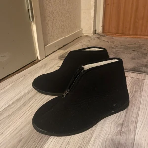 Svarta varmfodrade boots med dragkedja - Mysiga svarta ”uggs” med mjukt vitt foder och dragkedja framtill. Skorna har stilren design med platt sula och rund tå, perfekta för kalla dagar. Utsidan är i textil och insidan är fodrad för extra värme. Storlek 46
