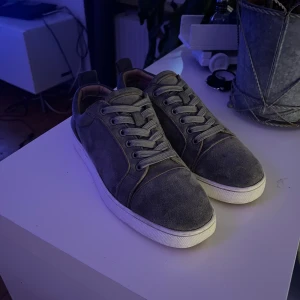 Grå mockasneakers från Louboutin - Snygga sneakers i grå mocka från Louboutin med klassisk röd sula och vita kanter. Skorna har rund tå, snörning och diskreta sömmar som ger en clean look. Perfekta för dig som gillar exklusiva detaljer och streetstyle.