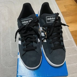 Adidas Campus 2 svarta sneakers - Adidas Campus 2 sneakers i svart mocka med vita detaljer och klassiska tre ränder på sidan. Skorna har rund tå, platt vit sula och snörning. Adidas-logga på plösen och hälen. Perfekt för dig som gillar stilrena och tidlösa sneakers.