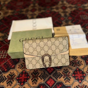 Gucci dionysus gg supreme mini bag shoulder - Snygg beige kuvertväska från Gucci med klassiskt GG-mönster över hela väskan. Väskan har en unik metallspänne framtill och insida i ljusbrunt skinn. Perfekt storlek för det viktigaste och en riktigt lyxig detalj till din outfit. Väskan är helt ny har inte används en enda gång. Priset kan diskuteras.