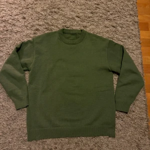 Olivgrön stickad tröja  - En stilren olivgrön stickad tröja med rund halsringning och ribbade muddar. Perfekt för lager-på-lager och passar till både jeans och chinos. Enkel design utan mönster eller logga, vilket gör den lätt att matcha med olika outfits.