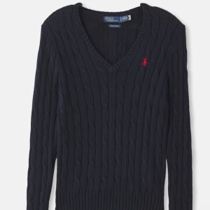 Marinblå kabelstickad tröja Polo Ralph Lauren - Kabelstickad marinblå tröja från Polo Ralph Lauren med röd broderad logga på bröstet. V-ringad modell med lång ärm och ribbade muddar. Snygg och klassisk passform, perfekt till jeans eller kjol. Ser helt ny ut och använd två gånger