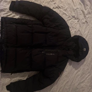  Sail Racing vinterjacka - Svart pufferjacka från Sail Racing med huva och dragkedja. Perfekt för kalla dagar och riktigt snygg streetstil. Lite skavanker, syns på bild men inget som förstör jackans funktion. Storlek L men passar även M
