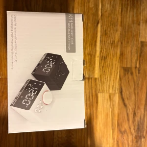 K11 Radio Alarm Clock with Dual USB Charger - Modern radio väckarklocka med stor digital display och dubbla USB-portar för laddning av iPhone, iPad och andra enheter. Finns i svart och vitt utförande. Produkten är ny i oöppnad förpackning.