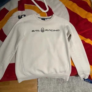 Vit Sail Racing sweatshirt i bomull - Vit sweatshirt från Sail Racing med svart logotyptryck på bröstet och liten logga i nacken. Klassisk rund halsringning och ribbade muddar vid ärmslut och nederkant. Tillverkad i mjuk bomull för en skön känsla. Perfekt för en clean och sportig stil.