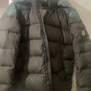 Moncler brun dunjacka med huva - Säljer en brun dunjacka från Moncler med glansiga gröna axelpartier och huva. Jackan har en quiltad design, dragkedja framtill och fickor på sidorna. Klassisk Moncler-logga på ärmen och snygg, puffig siluett som håller dig varm under kalla dagar.