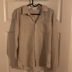 Beige linneskjorta  - Snygg beige skjorta i linne med klassisk krage och bröstficka. Skjortan har knappar framtill och långa ärmar, perfekt för en chill och stilren look. Passar dig som gillar enkel design.