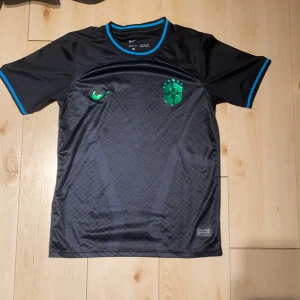 Svart Brasilien fotbollströja Nike - Snygg svart Brasilien fotbollströja från Nike med grönt Nike-märke och CBF-logga på bröstet. Tröjan har korta ärmar och blå detaljer vid krage och ärmslut. Tillverkad i lätt och ventilerande Dri-FIT-material med subtilt mönster på framsidan.