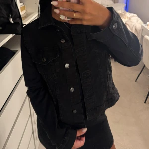 Svart jeansjacka med silvriga knappar - Snygg svart jeansjacka med klassisk krage och silvriga knappar framtill. Jackan har två bröstfickor med lock och knappar samt sidofickor. Rak passform och långa ärmar, perfekt att slänga över en hoodie eller t-shirt.