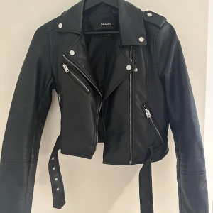 Svart skinnjacka från Pull&Bear - Snygg svart skinnjacka från Pull&Bear med klassisk bikerstil. Jackan har silvriga dragkedjor, knappar och bälte i midjan. Croppad modell med långa ärmar och  detaljer som axelklaffar och flera fickor med dragkedja. 