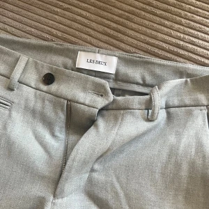 Ljusgrå kostymbyxor från Les Deux - Snygga ljusgrå kostymbyxor från Les Deux med klassisk passform och diskreta fickor. Byxorna har bälteshällor, knapp och dragkedja framtill. Perfekta för en stilren look och passar till skjorta eller t-shirt. OBS! Nypris 1800