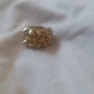 Guldig statementring med nitar - Cool och chunky ring i guldton med tre rader av pyramidformade nitar. Ringen har en bred design som verkligen sticker ut och ger en edgy vibe till din stil. Perfekt för dig som gillar att synas och vill ha något unikt på fingret. Storlek 18,5