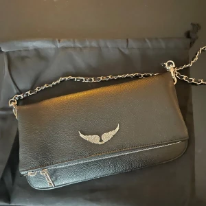 Svart axelväska från Zadig & Voltaire - Snygg svart axelväska i skinn från Zadig & Voltaire med silverfärgad kedja och ikonisk vingdetalj framtill. Väskan har en praktisk dragkedja och stilren design som passar perfekt till din outfit. Kommer med original dustbag.