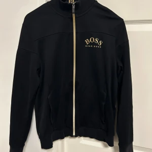 Svart ziptröja från Hugo Boss - Svart långärmad ziptröja från Hugo Boss med gulddetaljer. Stort BOSS-tryck på bröstet, guldfärgad dragkedja och dekorativa guldstickningar längs ena ärmen. Hög krage med Hugo Boss-logga. Perfekt för en clean och stilren look.