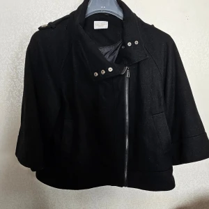 Svart ulljacka från Mac Scott, stl 40 - Stilren svart jacka från Mac Scott i ull med coola detaljer som sned dragkedja, knappar vid axlarna och trekvartsärmar. Jackan har hög krage och två fickor framtill. Perfekt för dig som gillar en edgy och modern look.