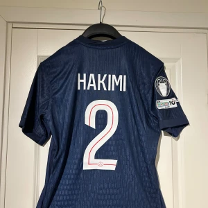 PSG Hakimi Champions League tröja - Helt ny Paris Saint-Germain matchtröja med Hakimi och nummer 2 på ryggen. Mörkblå med röd och vit rand framtill, Champions League-märke på ärmen och Qatar Airways som sponsor. Tillverkad i Dri-FIT material för bästa komfort. Klassisk Nike-logga och PSG-emblem på bröstet. Tröjan är en player version som gör den mycket skön att använda. Priset är inte hugget i sten!