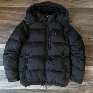 Svart pufferjacka från Polo Ralph Lauren - Svart pufferjacka från Polo Ralph Lauren med huva, flera dragkedjefickor och klassisk röd logga på bröstet. Jackan är vadderad och har en snygg, sportig look som passar perfekt när det är kallt ute. Performance-modell med detaljer i gult och blått på insidan.
