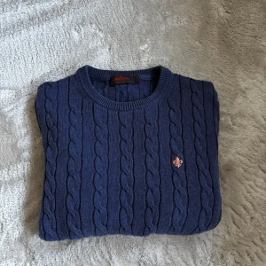 Marinblå kabelstickad tröja Morris - Snygg marinblå kabelstickad tröja från Morris i 100% pima bomull. Tröjan har rund hals, ribbade muddar och en orange broderad logga på bröstet. Perfekt för dig som gillar klassisk stil med en modern touch.