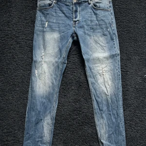 Blå baggy jeans med slitningar - Snygga blå baggy jeans med slitningar och ljusa tvättade partier på benen. Klassisk femficksmodell med dragkedja och knapp. Jeansen har en smal passform och är perfekta för en avslappnad streetstil. W36 L34.