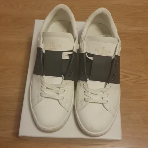 Valentino Garavani vita sneakers - Snygga vita sneakers från Valentino Garavani med bred svart rem över snörningen. Skorna har rund tå, platt sula och är tillverkade i skinn. Kommer med originalkartong, dustbag och äkthetskort. Perfekt för dig som gillar exklusiva och stilrena sneakers.