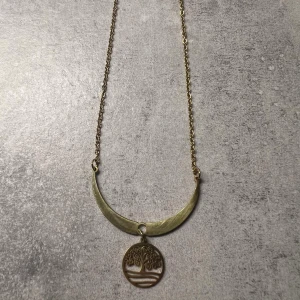 Guldigt halsband med livets träd - Unikt halsband i guldfärgad metall med tunn kedja och en halvmåneformad detalj. Hängsmycket är en rund platta med ett utskuret motiv av livets träd. Perfekt för dig som gillar boho-vibes och symboliska smycken. Lås med klassisk karbinhake.
