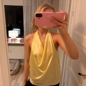 Gul omlott halterneck topp - Säljer en ljusgul halterneck topp med omlott-drapering framtill. Toppen är ärmlös och har öppen rygg, vilket ger en snygg och trendig look. Materialet är glansigt och känns mjukt mot huden, perfekt för varma dagar eller när du vill sticka ut.