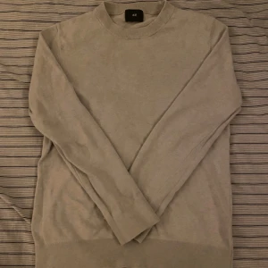 Ljusgrå stickad tröja från H&M - En enkel och stilren beige stickad tröja från H&M med rund hals och ribbade muddar. Perfekt för dig som gillar en clean look och vill ha något mjukt och bekvämt. Passar till jeans eller chinos och är lätt att matcha med andra plagg.