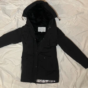 Svart parkajacka från Elvine - Svart parkajacka från Elvine med huva och flera fickor med dragkedja och knappar. Insidan har ett unikt foder med ugglemönster i svart och vitt. Jackan är lång, har snörning i midjan och är perfekt för kalla dagar. Materialet är polyester och jackan är långärmad.