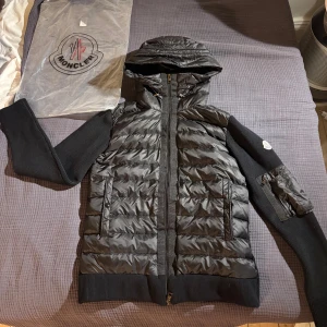 Svart Moncler cardigen med huva - Svart cardigan från Moncler med stickade ärmar och huva. Jackan har quiltad front och detaljer i blankt material, dragkedja fram och fickor med dragkedja. Moncler-logga på ärmen och extra ficka med dragkedja på ena armen. Snygg och modern look.