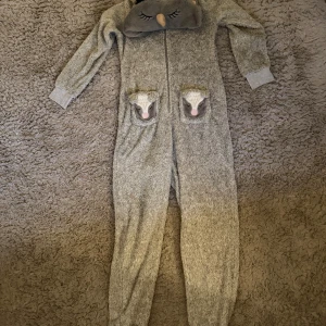 Grå jumpsuit med djurdetaljer från Hunkemöller - Supermysig grå jumpsuit från Hunkemöller med fluffigt material och dragkedja framtill. Huvan har söta broderade ögon och näbb, och fickorna pryds av gulliga djuransikten. Perfekt för chillkvällar hemma eller när du vill vara extra cozy.
