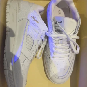 Axel Arigato sneakers - Säljer ett par stilrena vita sneakers från Axel Arigato. Skorna är tillverkade i premium skinn med snygga detaljer som guldlogga på sidan och präglad text på sulan. Klassisk rund tå, snörning och chunky sula ger en modern vibe. Perfekta för dig som gillar minimalistisk och clean stil. Skorna köptes i en pop up butik från Axel Arigato och är helt nya, aldrig använda. Be mig gärna om fler bilder. Nypris är 3750kr. Priset kan diskuteras.