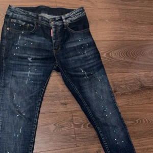 Dsquared2 blå jeans med färgstänk - Säljer ett par blå jeans från Dsquared2 med coola färgstänk och slitningar på benen. Jeansen har klassisk femficksdesign, orange kontrastsömmar och en snygg tvättad look. Passformen är rak med något smalare ben. Perfekta för dig som vill sticka ut lite extra.