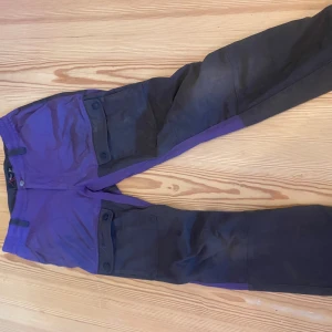 Lila och svarta friluftsbyxor High Mountain - Täckbyxor från High Mountain i lila och svart med stora fickor med knappar på benen. Byxorna har elastisk midja och är perfekta för utomhusaktiviteter. Snygg kontrast mellan färgerna och broderad logga på fickan.