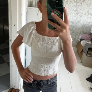 Vit smockad blus med volangärmar - Supersöt vit blus med smockad kropp och volangiga, korta ärmar. Toppen har fyrkantig ringning och kan bäras både offshoulder och på axlarna. Croppad modell med vågig kant nertill, perfekt till jeans eller kjol.